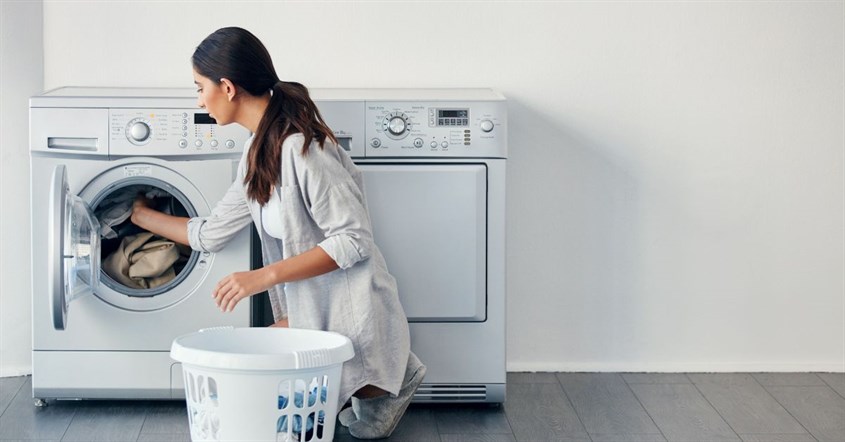  Wanita sedang memasukkan baju ke mesin cuci, sambil mempertimbangkan cara hemat listrik saat menggunakan alat elektronik.