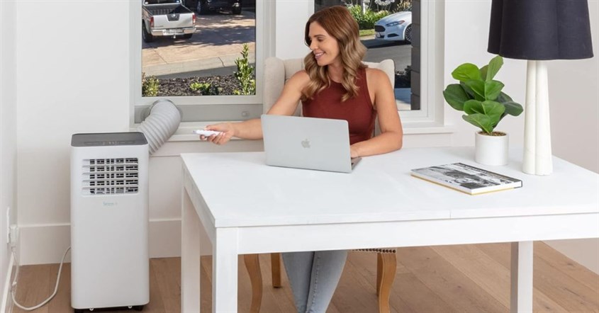 Seorang wanita bekerja di meja dengan laptop, menatap jendela, sambil mencari tips hemat listrik untuk perangkat elektronik.