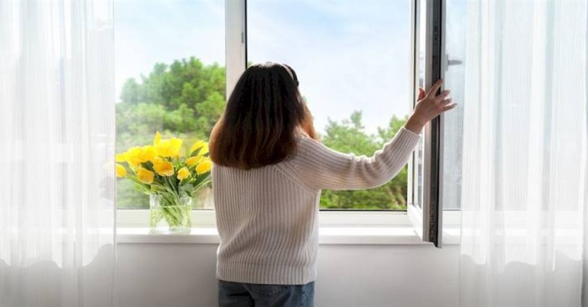 Seorang wanita berdiri di jendela, menikmati pemandangan sambil menerapkan pencahayaan alami untuk efisiensi energi.